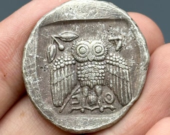 May include: Silver coin with an owl design. The owl is facing forward with its wings spread out. There are small details on the owl's feathers and eyes. The coin has a raised rim around the edge.