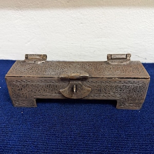 Könnte beinhalten: Eine rechteckige Box aus dunkel getöntem Metall mit aufwendigen, eingravierten kalligrafischen Mustern auf der gesamten Oberfläche. Die Box steht auf vier kurzen, integrierten Füßen und hat einen Klappdeckel mit einem zentralen Metallverschluss.