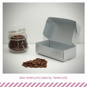 May include: A gray cardboard box template with a lid, open to reveal the inside. A glass jar filled with coffee beans sits on a white surface next to a pile of coffee beans.