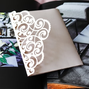 May include: A white, laser-cut, decorative envelope with a delicate, intricate design. The envelope is folded and placed on top of a closed book.