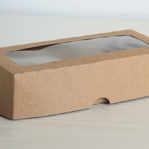 Puede incluir: Una caja de cartón marrón con una ventana de plástico transparente en la parte superior. La caja está diseñada para empaquetar y mostrar artículos pequeños.