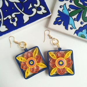 May include: A pair of square-shaped earrings with a blue border and a yellow, orange, and red paper flower design in the center.