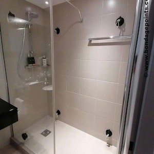 May include: A white tiled shower cubicle with a glass door and a chrome shower head. There are two black hooks on the wall inside the shower cubicle. There is a chrome towel rail on the wall outside the shower cubicle. The shower cubicle has a white tiled floor.