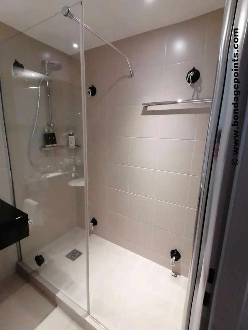 May include: A white tiled shower cubicle with a glass door and a chrome shower head. There are two black hooks on the wall inside the shower cubicle. There is a chrome towel rail on the wall outside the shower cubicle. The shower cubicle has a white tiled floor.