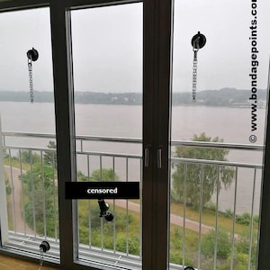 May include: Three silver chains with hooks attached to suction cups on a window. The chains are hanging down and are connected to handcuffs on the floor. The window overlooks a body of water and trees.