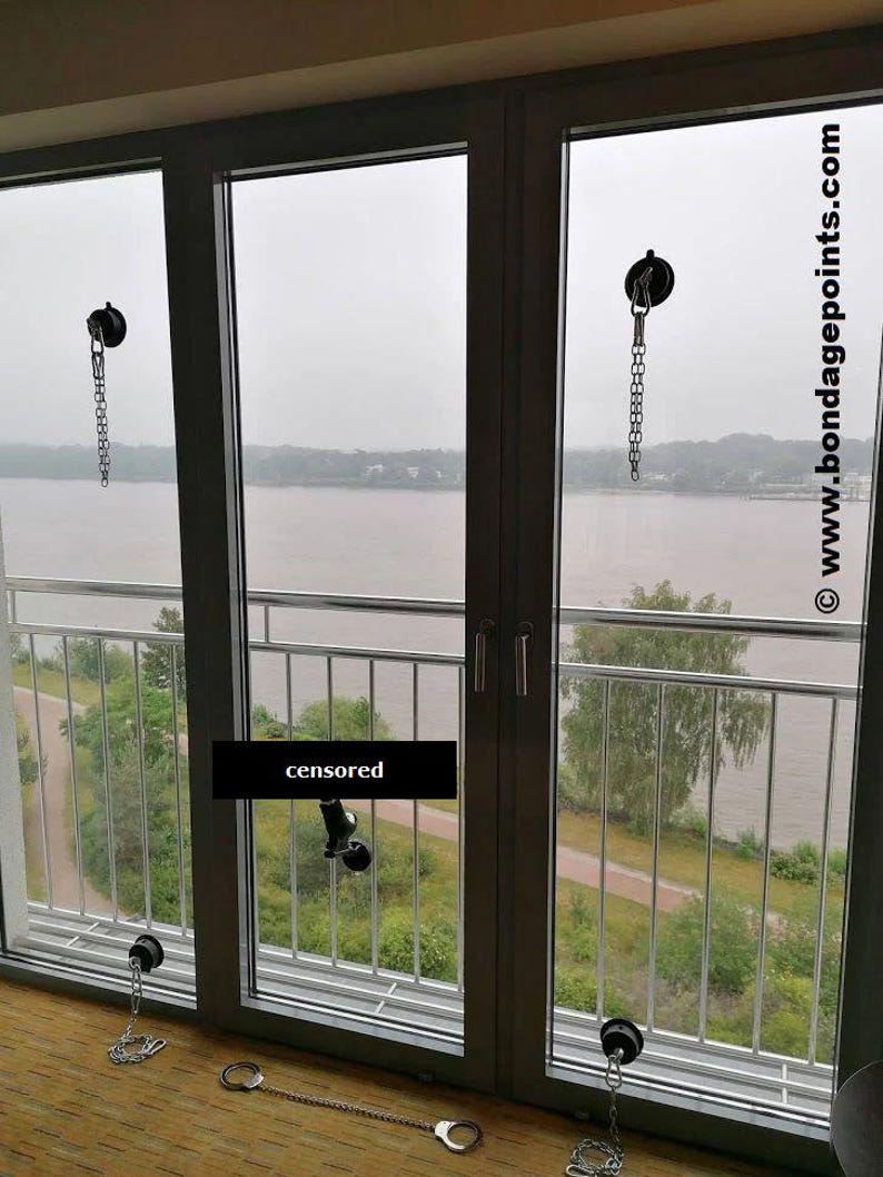 May include: Three silver chains with hooks attached to suction cups on a window. The chains are hanging down and are connected to handcuffs on the floor. The window overlooks a body of water and trees.