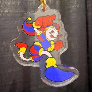 Puede incluir: Un llavero acrílico transparente con un personaje de dibujos animados en pose de carrera. El personaje tiene pelo rojo, un atuendo azul y rojo y detalles amarillos. El llavero tiene un anillo dorado.