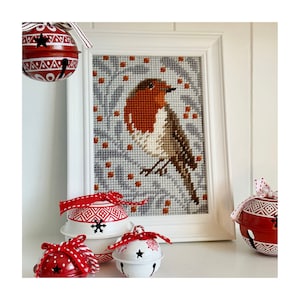 May include: A framed cross-stitch of a robin with a red breast, perched on a branch. The artwork is in a white frame. Decorative red and white jingle bells with star cutouts and red ribbons are in the foreground, suggesting a festive theme.