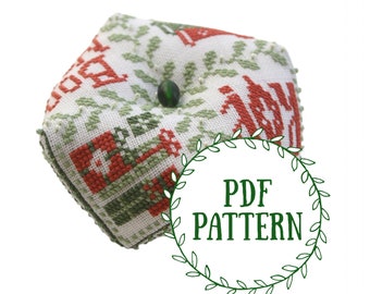 Op de afbeelding: Een groen en rood kruissteek kussen met een groene knoop in het midden. Het kussen is zeshoekig en heeft een witte achtergrond. De tekst "PDF PATTERN" staat in een groene cirkel met bladeren eromheen.