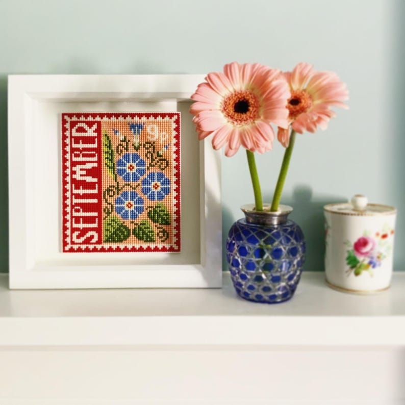 May include: A framed cross-stitch design featuring a red background with the word "SEPTEMBER" and a floral motif of blue flowers with green leaves. The frame is white and the flowers are in a blue and white vase.