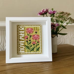 May include: A white frame with a yellow and green cross-stitch design of pink flowers. The design is labeled "NOVEMBER" and "11p".