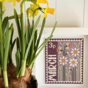 May include: A framed cross-stitch design of a purple stamp with white daisies and the text "3p MARCH".