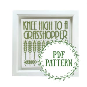 May include: A white frame with a green cross-stitch design that reads "Knee High to a Grasshopper". The design also includes a row of wheat stalks. A green wreath with the text "PDF PATTERN" is in the foreground.