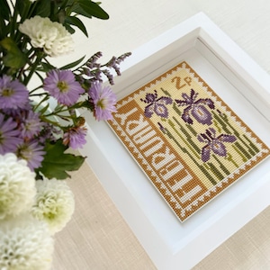 May include: A framed cross-stitch piece depicting a vintage postage stamp with purple irises and the text "The Iris" and "2p".