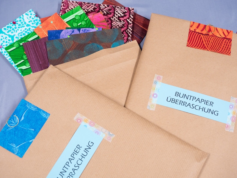 May include: Two brown paper envelopes containing assorted colorful patterned papers. Each envelope features a light blue label with the text BUNTPAPIER &Uuml;BERRASCHUNG, secured with decorative tape. The papers inside display various floral and geometric designs.