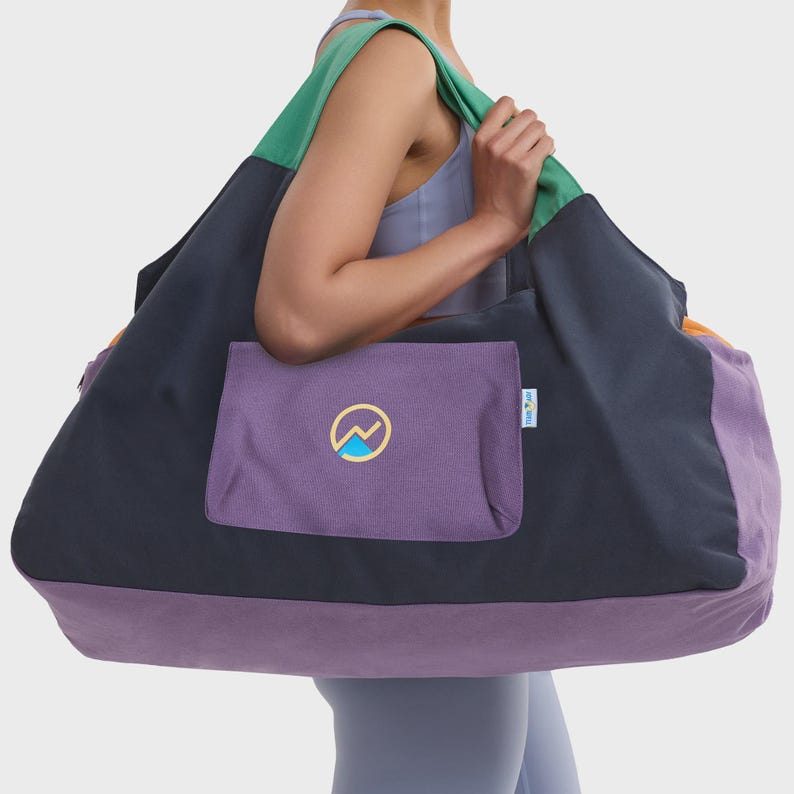 May include: A large color-blocked duffel bag featuring a dark charcoal main body, a purple base, and green shoulder straps. A purple exterior pocket displays a circular logo with a yellow outline and a blue mountain graphic. A small white tag is visible.