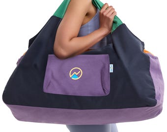 May include: A large color-blocked tote bag featuring a dark navy main body, a purple front pocket, and green shoulder straps. The front pocket displays a circular logo with a yellow outline and a blue mountain graphic. An orange side panel is visible.