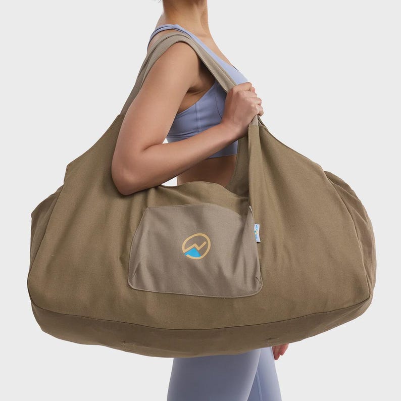 May include: A large, olive-green canvas hobo-style bag featuring a wide shoulder strap and a central exterior pocket. The pocket displays a circular logo with a blue mountain graphic and a yellow outline. A small white fabric tag is visible on the side.