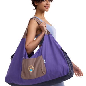 May include: A large hobo-style shoulder bag featuring a vibrant purple main body, a dark navy blue bottom panel, and a taupe-colored shoulder strap. A taupe exterior pocket with a zipper closure displays a circular logo with a mountain graphic.