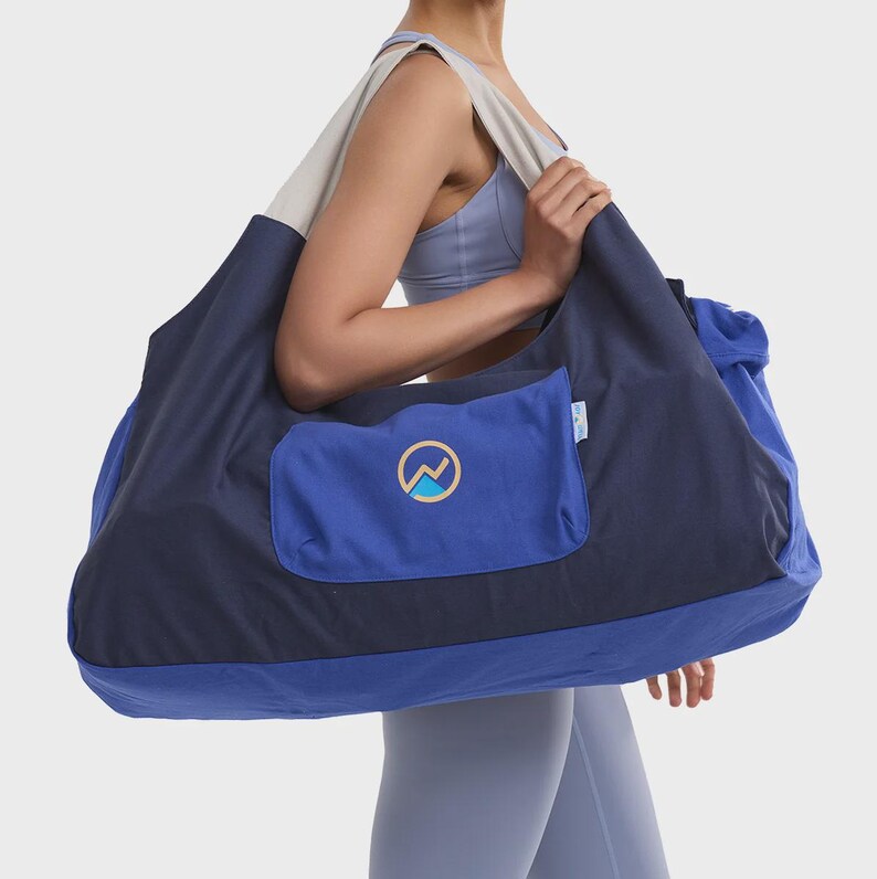 May include: A large, multi-toned blue and navy shoulder bag featuring a wide, light-colored strap and a rectangular front pocket. The pocket displays a circular logo with a stylized mountain graphic in yellow and light blue, with a small side tag.