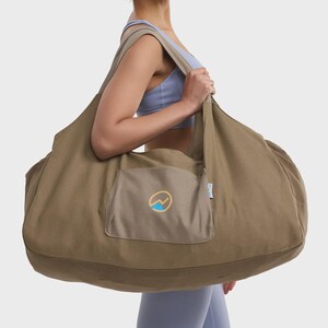 May include: A large, olive-green canvas hobo-style bag featuring a wide shoulder strap and a central exterior pocket. The pocket displays a circular logo with a stylized mountain peak in yellow and blue. The bag has a soft, unstructured shape.