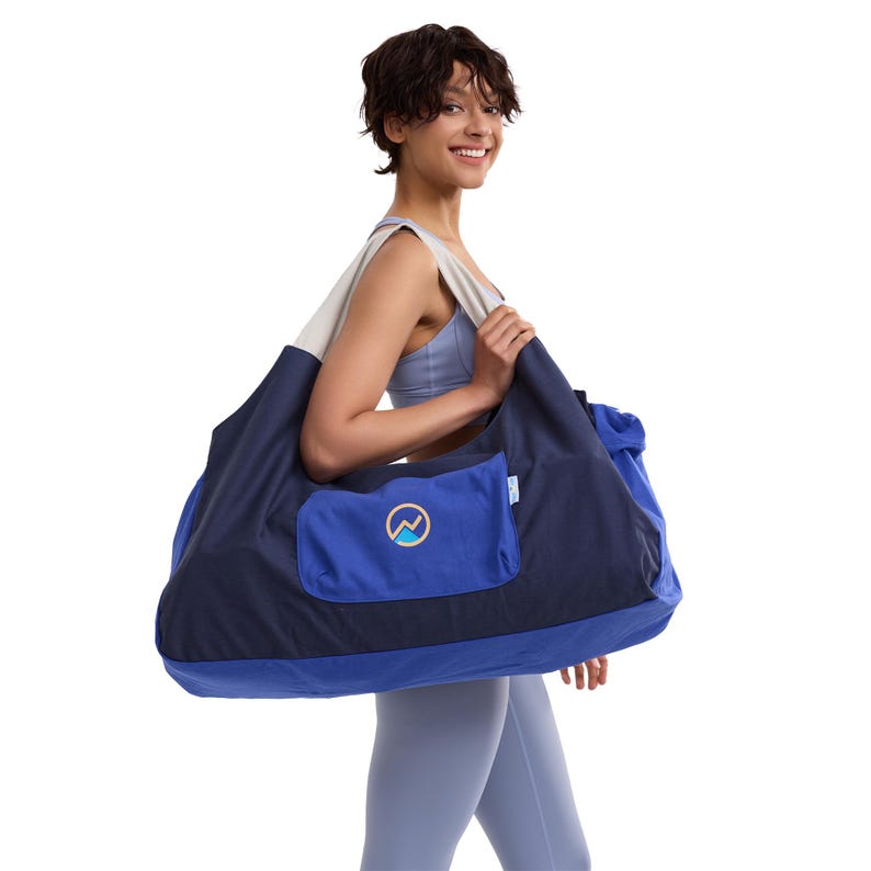 May include: A large duffel bag featuring a dark navy blue upper section and a vibrant royal blue lower section. The bag includes a wide light gray shoulder strap and a rectangular front pocket with a circular logo containing a mountain graphic.