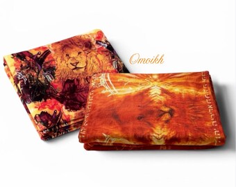 May include: Two folded blankets featuring vibrant lion imagery. One blanket displays a detailed lion face with warm orange and red tones, while the other shows a lion surrounded by abstract floral patterns. The text Omoikh is visible.