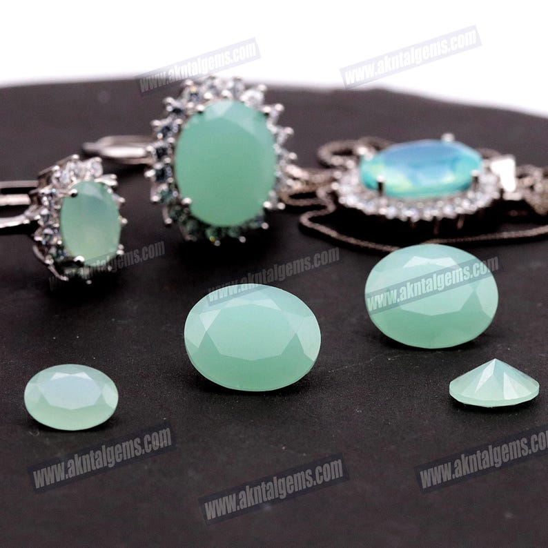 Op de afbeelding: Een assortiment lichtgroene edelstenen en sieraden. De afbeelding toont twee ringen, een hanger en verschillende losse, ovaal geslepen edelstenen. De sieraden hebben zilveren zettingen. De edelstenen zijn facetgeslepen en de achtergrond is zwart.
