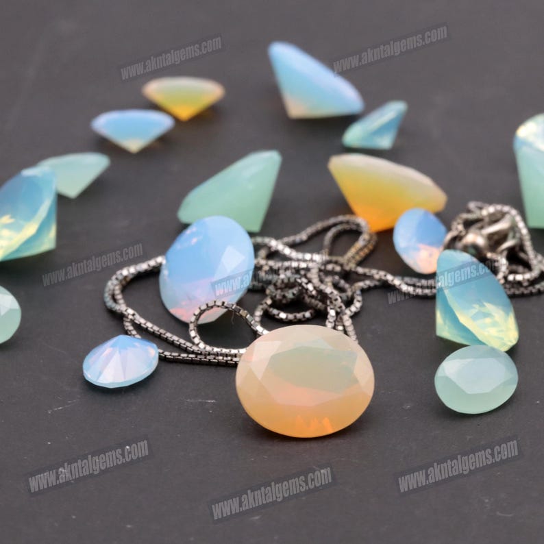 Op de afbeelding: Een assortiment opalescente edelstenen in verschillende vormen en maten, waaronder ovale, druppel- en driehoekige slijpsels. Een zilveren ketting is tussen de edelstenen gedrapeerd. De edelstenen vertonen een scala aan kleuren, van lichtblauw tot geel.
