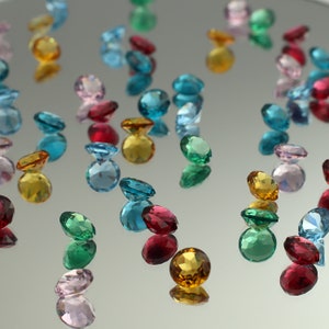 Puede incluir: Surtido de piedras preciosas facetadas de colores en varios tonos de azul, verde, rojo, rosa y amarillo. Las piedras preciosas están dispersas sobre una superficie reflectante.