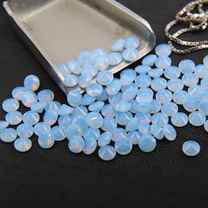 May include: A large quantity of light blue opal gemstones, scattered on a black surface. The gemstones are round and have a faceted cut.