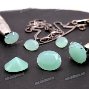 Op de afbeelding: Een verzameling lichtgroene edelstenen in verschillende vormen, waaronder ovale, ronde en facetgeslepen stenen. Een zilveren ketting en sieraden gereedschap zijn ook aanwezig. De edelstenen worden op een donkere ondergrond getoond.