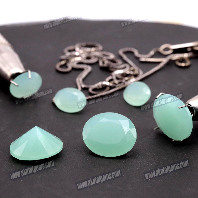 Op de afbeelding: Een verzameling lichtgroene edelstenen in verschillende vormen, waaronder ovale, ronde en facetgeslepen stenen. Een zilveren ketting en sieraden gereedschap zijn ook aanwezig. De edelstenen worden op een donkere ondergrond getoond.
