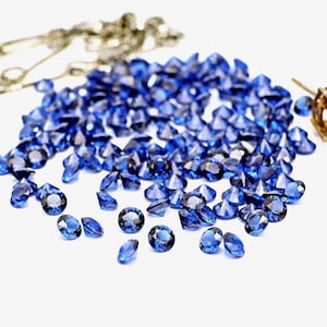May include: A collection of brilliant blue gemstones scattered on a white surface. The gems vary in size and shape, with facets catching the light. A gold chain and earring are visible in the background.