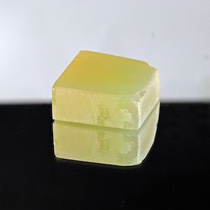 Puede incluir: Un bloque rectangular transl&uacute;cido de color amarillo verdoso con una superficie rugosa y texturizada. El jab&oacute;n est&aacute; sobre una superficie negra y refleja la luz.