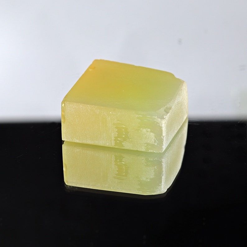 Puede incluir: Un bloque rectangular transl&uacute;cido de color amarillo verdoso con una superficie rugosa y texturizada. El jab&oacute;n est&aacute; sobre una superficie negra y refleja la luz.