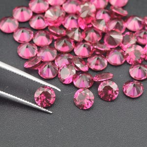 May include: A close-up of a group of round, faceted, pink gemstones. The gemstones are scattered on a black surface. One gemstone is being held by a pair of tweezers.