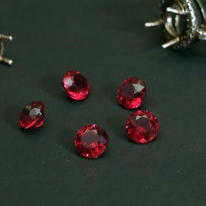 Puede incluir: Cinco gemas facetadas de color rojo intenso se encuentran sobre una superficie oscura. Las gemas varían en forma, con cortes redondos y ovalados. En el fondo, se aprecia un engaste de plata para joyería, sugiriendo piedras sueltas para crear joyas.