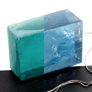 Puede incluir: Un colgante rectangular con un degradado de color de verde azulado a azul. El colgante tiene una superficie texturizada y se muestra con una cadena de plata. El colgante probablemente está hecho de resina o un material similar.