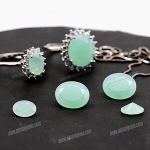 Op de afbeelding: Een verzameling lichtgroene edelsteen sieraden en losse stenen. De afbeelding toont twee ringen, een ketting en verschillende ovale en facetgeslepen edelstenen. De sieraden hebben een zilveren zetting. De edelstenen worden op een donkere ondergrond getoond.