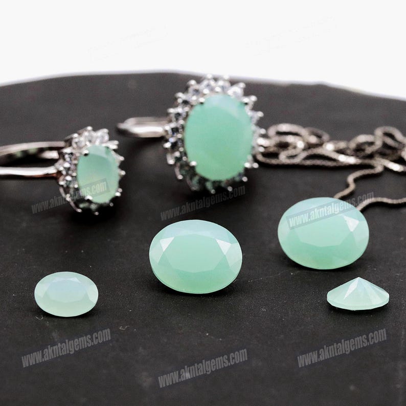 Op de afbeelding: Een verzameling lichtgroene edelsteen sieraden en losse stenen. De afbeelding toont twee ringen, een ketting en verschillende ovale en facetgeslepen edelstenen. De sieraden hebben een zilveren zetting. De edelstenen worden op een donkere ondergrond getoond.
