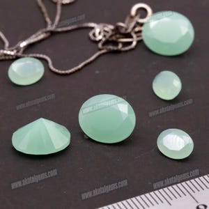 Op de afbeelding: Een verzameling gefacetteerde, lichtgroene edelstenen in verschillende vormen en maten, waaronder ovaal en rond. Een zilveren ketting is gedeeltelijk zichtbaar op de achtergrond. De edelstenen worden op een donkere ondergrond gepresenteerd.