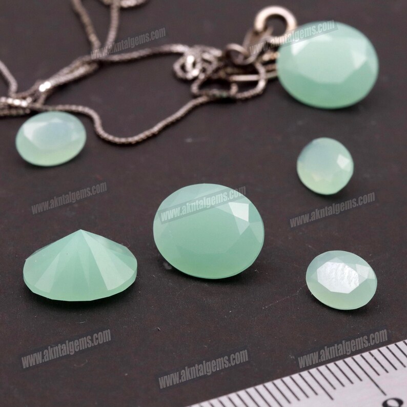 Op de afbeelding: Een verzameling gefacetteerde, lichtgroene edelstenen in verschillende vormen en maten, waaronder ovaal en rond. Een zilveren ketting is gedeeltelijk zichtbaar op de achtergrond. De edelstenen worden op een donkere ondergrond gepresenteerd.