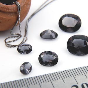 May include: An assortment of faceted, oval-shaped, dark gray gemstones. One gemstone is set in a silver chain necklace. A ruler is in the background. The gemstones vary in size, with a few larger stones and several smaller ones.