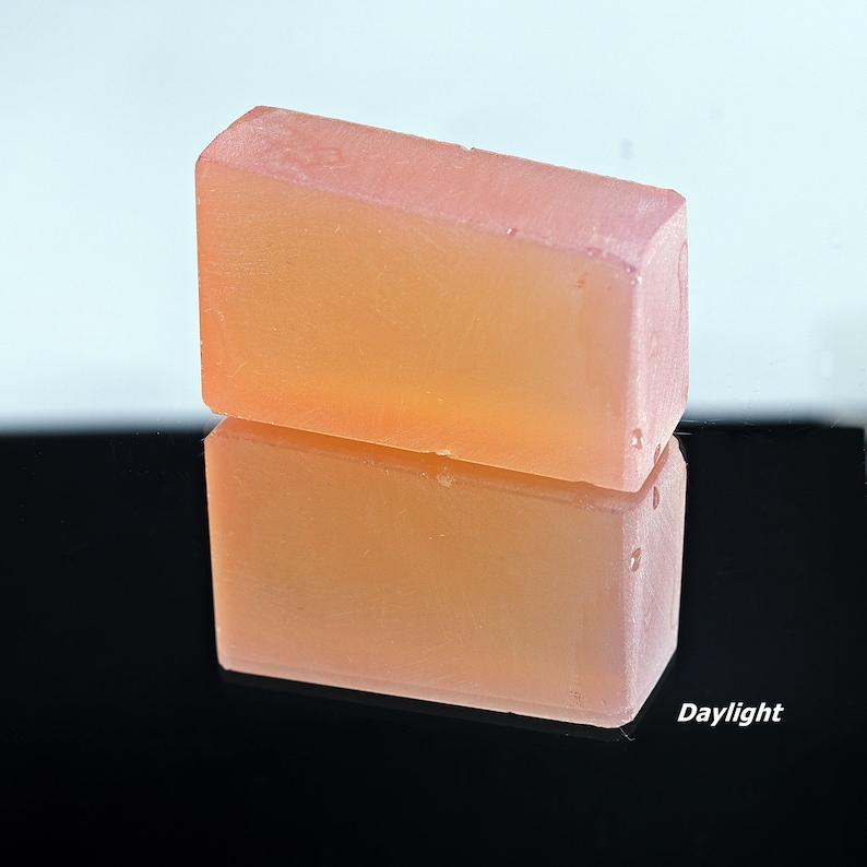 Puede incluir: Una barra de jab&oacute;n rectangular transl&uacute;cida con un tono rosa claro. El jab&oacute;n se refleja en una superficie negra.
