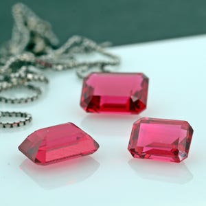 Peut inclure: Trois pierres précieuses rouges taillées en émeraude sur une surface blanche. Les pierres précieuses sont facettées et ont un fort éclat.