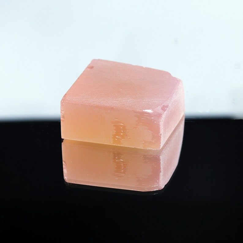 Puede incluir: Una barra de jab&oacute;n rectangular de color rosa p&aacute;lido con un aspecto ligeramente esmerilado. El jab&oacute;n est&aacute; sobre una superficie negra y se refleja en la superficie.