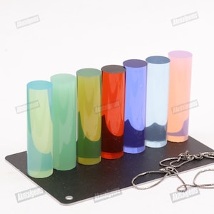 May include: A collection of seven colorful, cylindrical gemstones in shades of blue, green, yellow, red, purple, and pink. The gemstones are arranged on a black rectangular base, with a silver chain in the foreground. The gemstones are translucent.