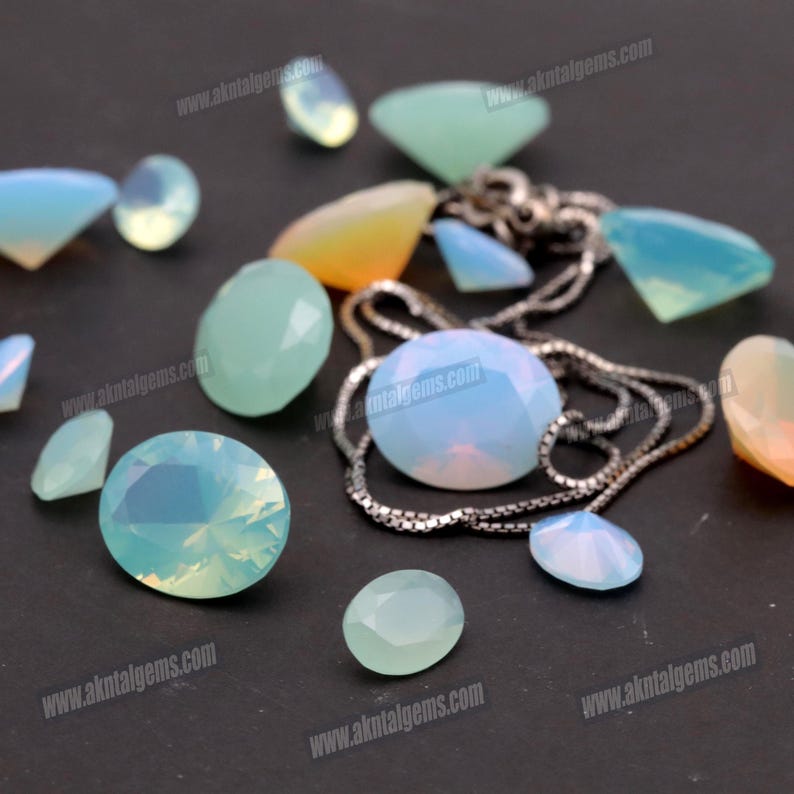 Op de afbeelding: Assortiment van opalescente edelstenen in diverse vormen, waaronder rond, ovaal en druppelvormig. Een zilveren ketting is verweven tussen de edelstenen. De edelstenen vertonen een scala aan kleuren, van lichtblauw tot geel en groen, met een glinsterend effect.