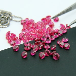 May include: A close-up of a pile of pink gemstones, some of which are being held by a pair of tweezers. The gemstones are scattered on a black surface.